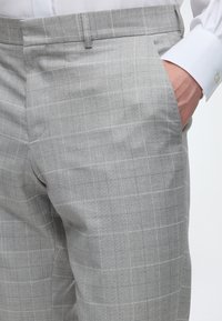 Personne portant un pantalon à carreaux gris clair et une chemise blanche à manches longues, une main dans la poche.