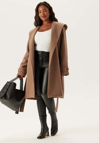 Femme portant un manteau camel, un haut blanc, un pantalon en cuir noir taille haute, des bottes à talons noires, tenant un grand sac à main noir dans la main droite.