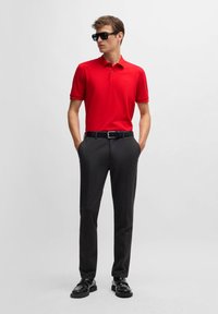 Röd pikétröja, svarta byxor, svart bälte och glänsande svarta skor. Korta ärmar, enfärgade med en diskret logotypdetalj. Slät textur.