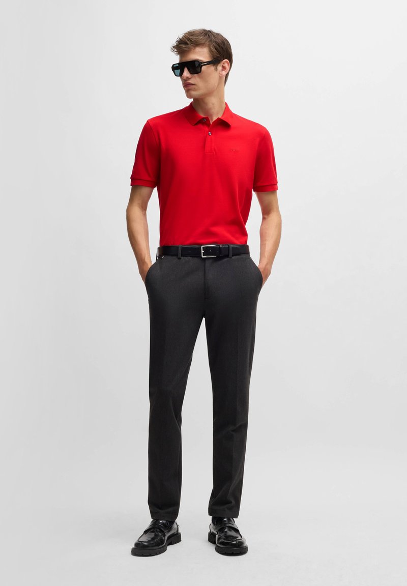 Röd pikétröja, svarta byxor, svart bälte och glänsande svarta skor. Korta ärmar, enfärgade med en diskret logotypdetalj. Slät textur.