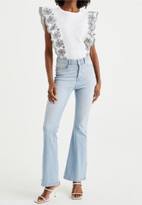 Vit T-shirt med svart blommönsterbroderi och volangärmar, kombinerad med ljusblå utsvängda jeans och vita sandaler med remmar.