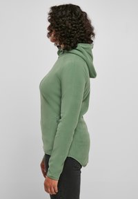 Felpa in pile verde con design aderente, cappuccio e tasca laterale con zip. Texture liscia, maniche lunghe e orlo leggermente curvo.
