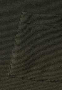 Tmavě olivově zelená látka s texturovaným, nařaseným povrchem. Obsahuje čtvercovou kapsu přišitou na materiál. Švy jsou čisté a jednoduché.