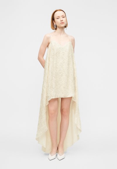 Φόρεμα σε κρεμ απόχρωση με υφή, λαιμόκοψη σε σχήμα V και ασύμμετρο τελείωμα. Ρυθμιζόμενες λεπτές τιράντες, συνδυασμένο με λευκά παπούτσια μυτερά στις μύτες.