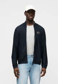 Μπλε μπουφάν με φερμουάρ και υφή υφάσματος, με ελαστικές μανσέτες, φορεμένο πάνω από γκρι ανοιχτό μπλουζάκι, συνδυασμένο με ανοιχτό μπλε τζιν. Άσπρο καπέλο.