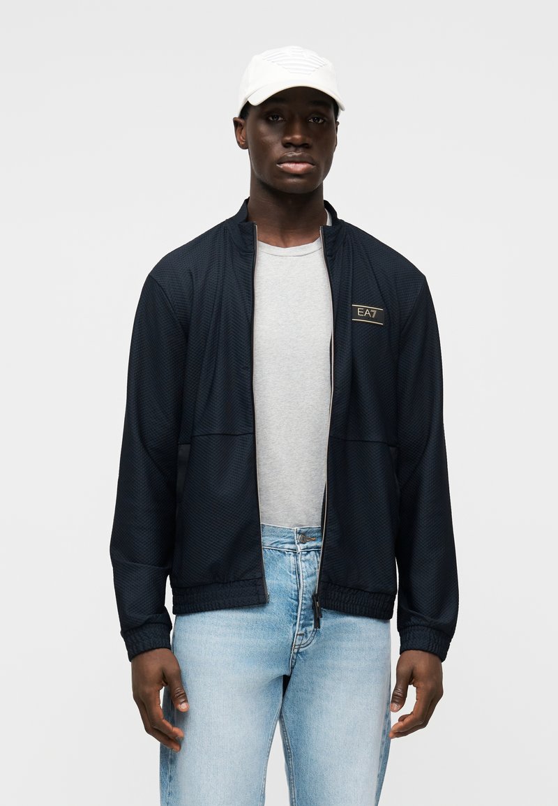 Μπλε μπουφάν με φερμουάρ και υφή υφάσματος, με ελαστικές μανσέτες, φορεμένο πάνω από γκρι ανοιχτό μπλουζάκι, συνδυασμένο με ανοιχτό μπλε τζιν. Άσπρο καπέλο.