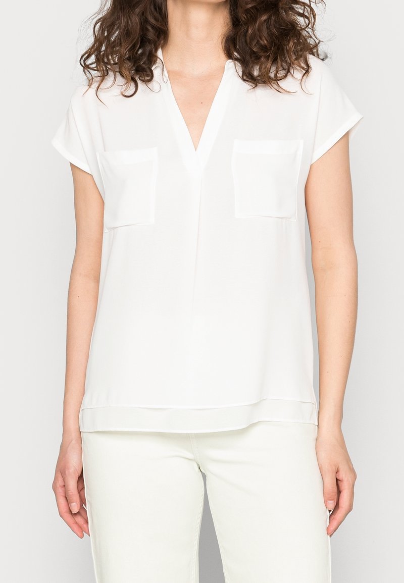 Femme portant un chemisier blanc à manches courtes avec deux poches sur le devant et un pantalon blanc, sur un fond clair uni.
