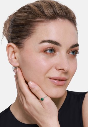 Bague en argent avec une pierre ronde en malachite verte, bande texturée, portée sur une main avec des ongles naturels, sur fond uni.