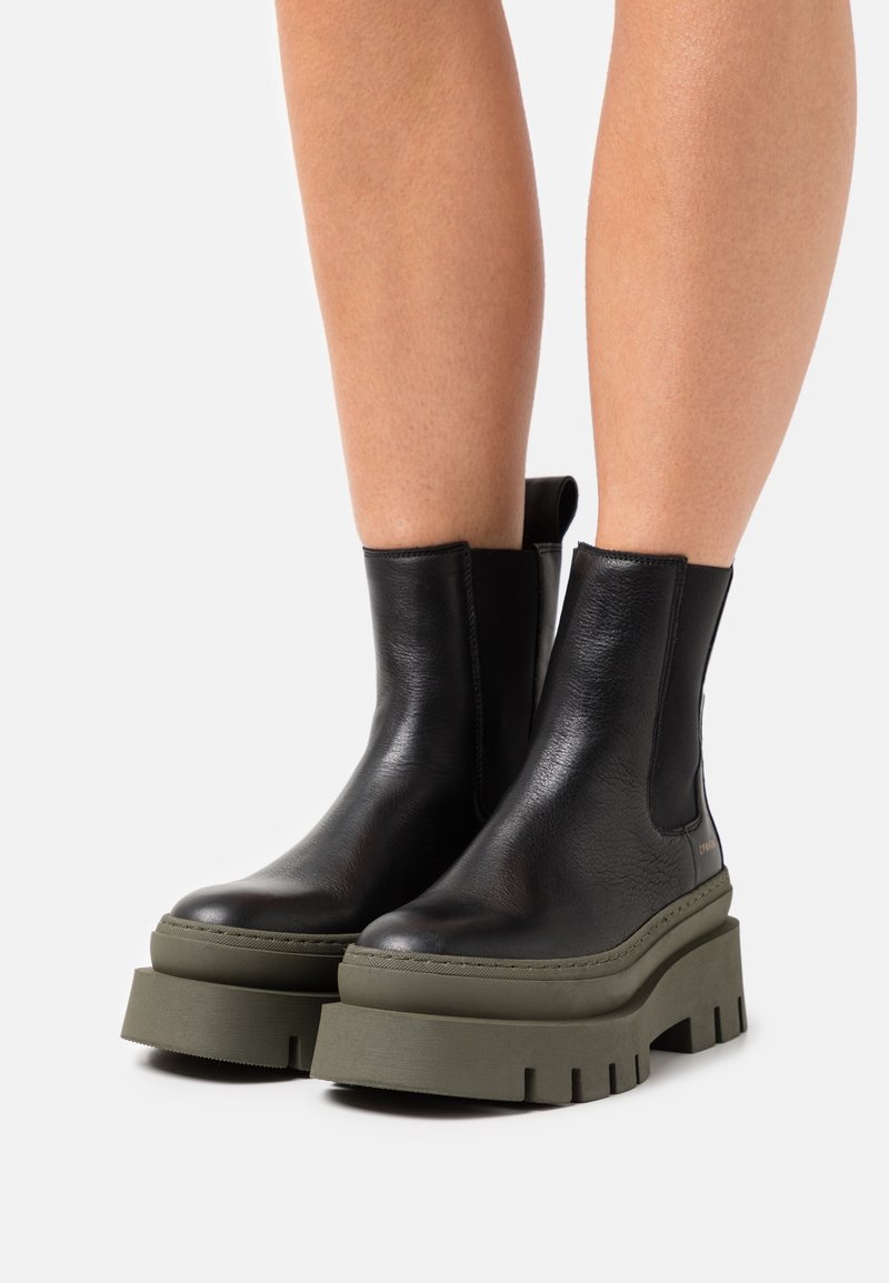 Jambes portant des bottines en cuir noir à talons épais avec semelles compensées vertes sur fond blanc.