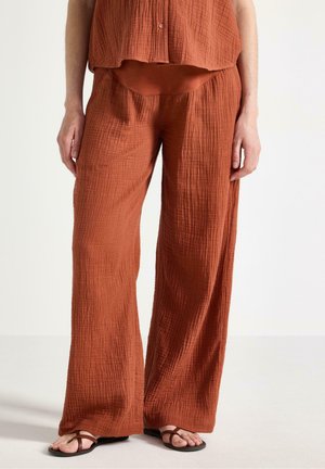 Person iført løstsiddende, teksturerede, rustfarvede vidde ben-bukser og matchende knaplukket top, parret med brune flade sandaler på hvidt gulv.