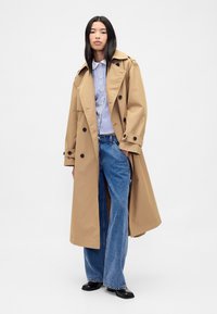 Beige trenchcoat i slät tyg med dubbelknäppning, stora svarta knappar och stora slag. Kombinerad med en randig skjorta och vida jeans.