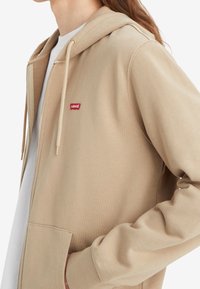 Beige zip-up hoodie tillverkad av mjukt tyg, med en framficka, dragskoHuva och en liten röd Levi's-logotyp på bröstet.
