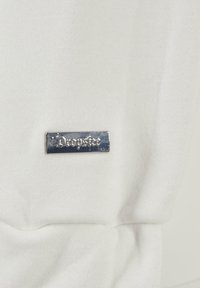 Tissu blanc avec une texture lisse, présentant une étiquette en métal argenté embossée avec "Dropske". L'étiquette est rectangulaire avec des coins arrondis.