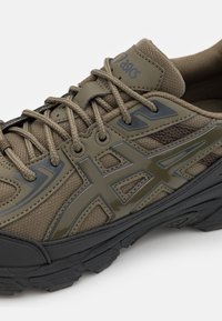 Shoe atlético verde oliva com parte superior em malha, atacadores e sola de borracha escura. Apresenta um logotipo brilhante e realces, design texturizado e materiais duráveis.