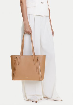 Mujer con pantalones blancos de pierna ancha y chaleco sostiene un bolso tote de cuero marrón claro con doble asa y detalles de cremallera en la parte frontal.