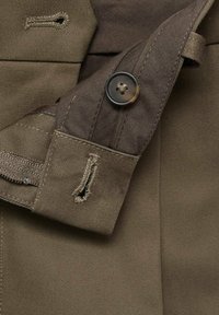 Brown fabric detail showing a button, stitched edges, and a small slit. Texture is smooth with a slight sheen and diagonal weave pattern.