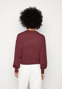 Osoba z kręconymi włosami, zwrócona plecami, ubrana w bordowy sweter z długim rękawem i białe spodnie, na jednolitym białym tle.