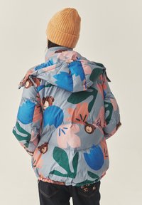 Puffervest i lyseblå med farverige blomster- og bjørne mønstre, med en pelsforet hætte og knapdetaljer. Båret med mørkeblå jeans.