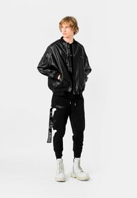 Chaqueta bomber de imitación de cuero negra con puños acanalados, combinada con pantalones joggers negros que presentan un parche gráfico y cremalleras. Botas de combate blancas.