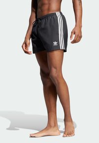 Svarte svømmebukser med hvitt Adidas-logo og tre vertikale striper på sidene. Lett stoff, elastisk midjebånd med snor.