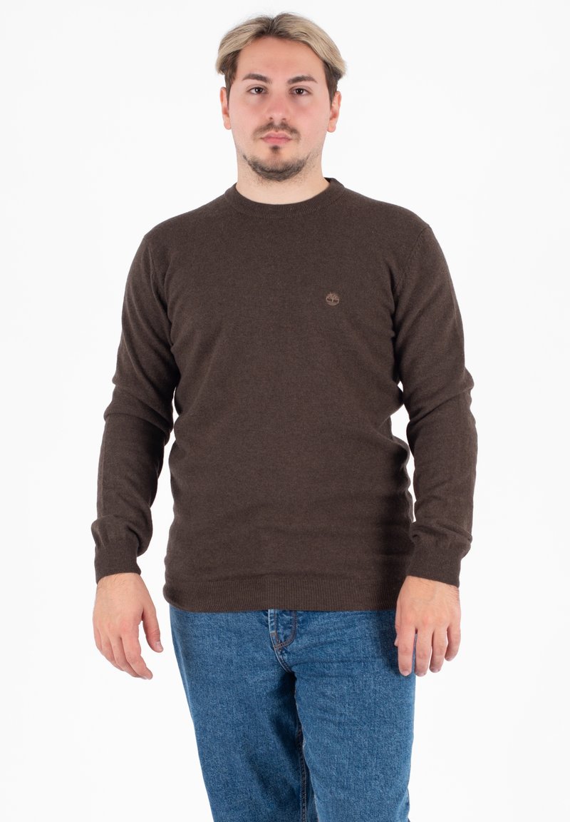 Maglione marrone a maniche lunghe con scollatura rotonda e polsini a coste, caratterizzato da un piccolo logo sul petto. Abbinato a jeans blu.
