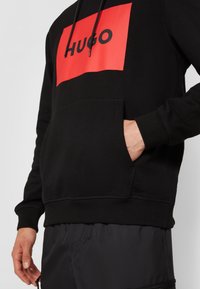 Sweat à capuche noir avec un logo rectangulaire rouge affichant "HUGO" en lettres noires grasses, doté d'une poche avant et de poignets et ourlet côtelés.