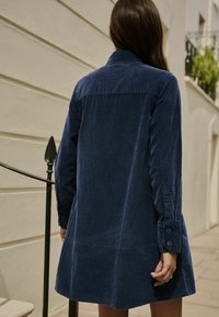 Granatowa sukienka koszulowa z sztruksu z kołnierzykiem, długimi rękawami i guzikami przy mankietach, charakteryzująca się rozkloszowanym krojem i przeszyciami.