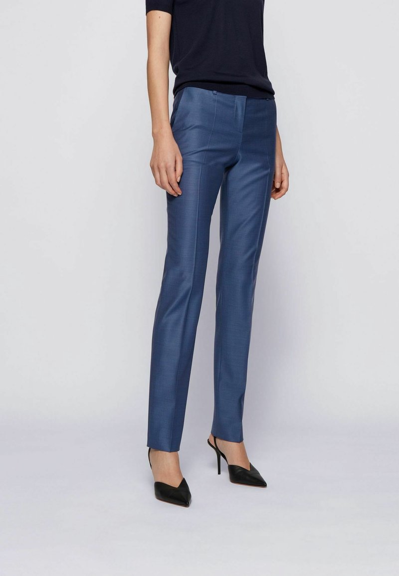 Personne portant un pantalon de costume bleu slim, des chaussures à talons noires et un haut noir à manches courtes, debout devant un fond clair uni.