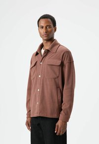 Bruine fleecejas met een spitse kraag, twee borstzakken en zilverkleurige drukknoopsluitingen. Gecombineerd met een lichtgekleurde blouse en donkere broek.