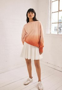 Różowo-pomarańczowy sweter w ombre z mankietami, zestawiony z białą plisowaną spódnicą i jasnymi trampkami na drewnianej podłodze.