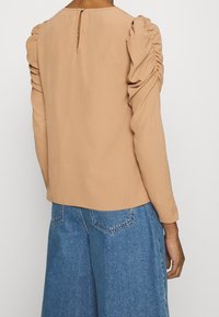 Maglia a maniche lunghe in tessuto beige chiaro con maniche a palloncino arricciate. La schiena presenta un piccolo apertura a chiave e una cerniera nascosta. Fit casual.