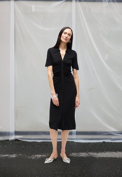 Crna haljina na kopčanje s kratkim rukavima, naboranim detaljima i ovratnikom. Kombinirana sa srebrnim balerinama i prozirnom narukvicom. Jednostavan i utegnut kroj.