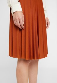 Plisovaná oranžová sukně vyrobená z texturované látky. Detailní záběr ukazuje lem a ruku položenou na látce, což zdůrazňuje plisé.