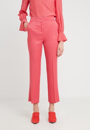 Vrouw draagt koraalroze op maat gemaakte broek met een bijpassende blouse met lange mouwen en rode mules met puntige neus, staand tegen een effen achtergrond.