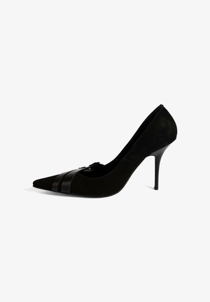 Chaussure noire à talon haut avec un bout pointu, en daim. Elle présente une sangle noire brillante à l'avant et un talon aiguille élancé.