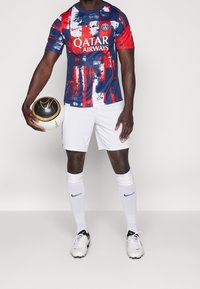 Fotbalový dres s krátkým rukávem v námořnické modré a červené barvě s abstraktním malířským vzorem, obsahující logo "QATAR AIRWAYS", bílé šortky a černobílý fotbal.