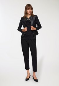 Giacca in pelle sintetica nera con un design aderente, chiusura con zip e colletto rialzato, abbinata a una maglietta nera e pantaloni neri a sigaretta.