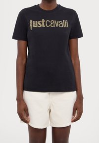 Czarna bawełniana koszulka z krótkim rękawem, z metalicznym złotym napisem "justcavalli" z przodu. Okrągły dekolt i luźny fason. W połączeniu z kremowymi szortami.