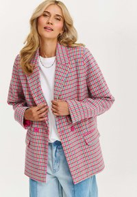 Blonďatá žena v růžovém, modrém a bílém houndstooth saku s růžovými knoflíky, bílém tričku, modrých džínách a barevném korálkovém náhrdelníku.