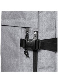 Grå tygryggsäck med en texturerad yta, utrustad med svarta dragkedjor, ett spännlock och en förstärkt rem för säker fastsättning.