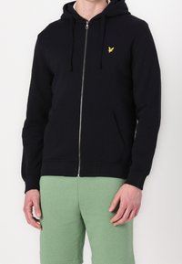 Muž oblečený v černé mikině na zip s malým žlutým ptáčkem na hrudi a v zelených šortkách, stojící na bílém pozadí.