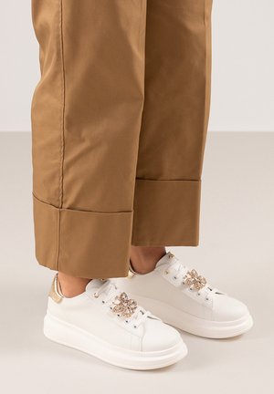 Zapatillas blancas con un acento dorado brillante, suela texturizada y detalle decorativo de strass; combinadas con pantalones cortos color marrón claro.