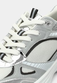 Ανοιχτό γκρι αθλητικό παπούτσι με διαφανές υλικό, λευκές κορδόνες, μεταλλικά ασημένια στοιχεία και μαύρες ρίγες. Τρύπες αερισμού στα πλάγια.