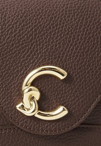 Brown textured leather handbag featuring a prominent gold-tone hardware logo detail. Includes a curved and twisted design element.