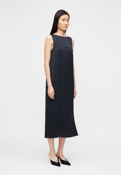 Varrukateta tumesinist värvi sirge lõikega kleit, mis ulatub pahkluuni. Sile tekstuur, minimaalsed õmblused, külje lõhikud, sobitatud mustade teravate madalate kingadega.