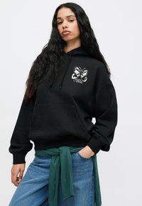 Fekete kapucnis pulóver, nagy fehér pillangó grafikával, kenguru zsebbel és állítható húzózsinórral. Kék farmernadrág fölött viselve, zöld kötővel.