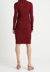 Robe bordeaux côtelée à manches longues avec une silhouette ajustée, s'arrêtant au milieu du mollet. Accompagnée de talons ouverts beige.