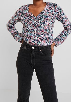 Personne portant un chemisier à manches longues à fleurs roses, blanches et bleu sarcelle, associé à un jean noir taille haute sur un fond uni.
