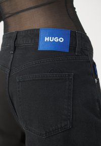 Jean en denim noir avec un patch en cuir bleu portant l'inscription "HUGO" sur la ceinture et un détail de poche arrière, présentant un motif texturé subtil.