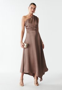 Abito in satin marrone con design a una spalla, orlo asimmetrico fluido e texture liscia. Indossato con accessori dorati e sandali.